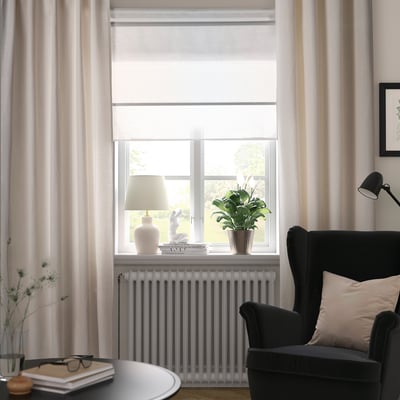 Des rideaux LENDA blanc cassé à tissage linéaire rehaussent les fenêtres hautes. Fauteuil noir à l’échelle.