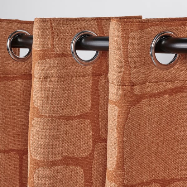 Deux rideaux orange brûlé pendent à des œillets d’argent sur une tringle sombre, bloquant la lumière, avec un motif tissé en jacquard.