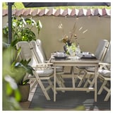 Ensemble de salle à manger d’extérieur avec chaises beiges, table blanche, tapis noir et blanc, plantes en pot et vaisselle sous auvent.