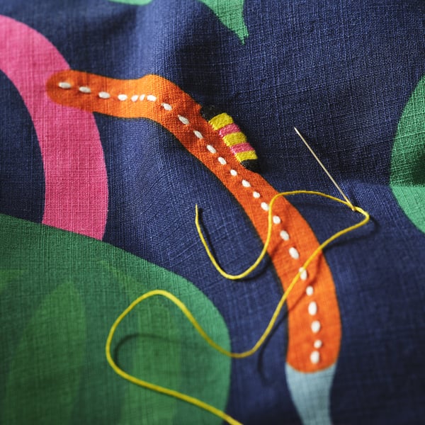 Gros plan d’une housse de coussin brodée à la main avec une chenille orange et un motif abstrait coloré, mettant en valeur des coutures complexes et des couleurs vives.