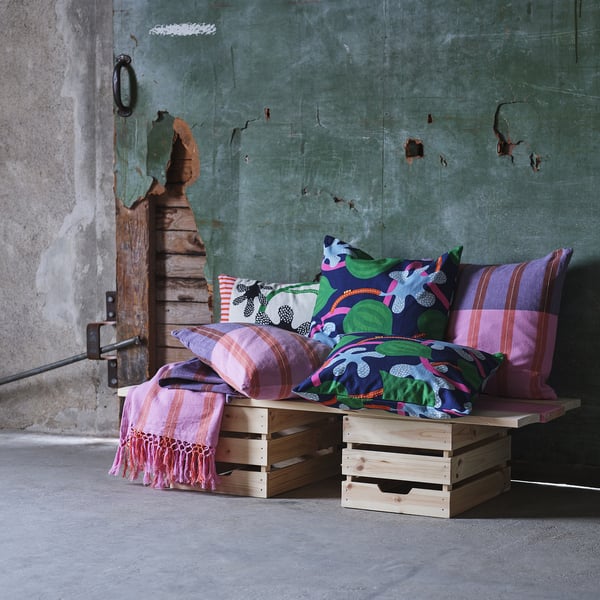 Coussins colorés avec des motifs sur des caisses en bois contre un mur rustique. Les nuances de violet, de vert, de rose et de bleu créent une ambiance animée et chaleureuse.