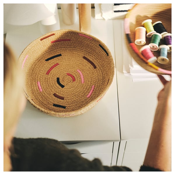 Imaginez un panier rond en jute tressé avec des détails brodés colorés. Le panier repose sur une surface blanche avec des bobines de fil à côté.