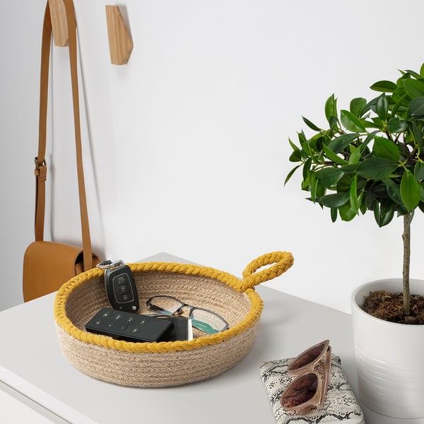 Le panier en jute à bords jaunes sur la table contient des clés, une télécommande et des verres. Un sac à main brun est suspendu à côté. Une plante en pot se trouve à proximité.