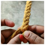 Des mains fabriquent un panier en jute MÄVINN jaune moutarde avec du vernis à ongles rouge.