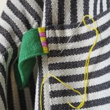 Gros plan d’un tablier en tricot rayé noir et blanc avec une bretelle verte, du fil jaune et une aiguille ; mettant en valeur l’artisanat détaillé.