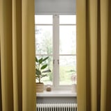 Fenêtre avec rideaux MAJGULL beige-jaune ouverts, mettant en valeur la texture douce et l’intimité, plante, bougie et radiateur blanc en dessous.