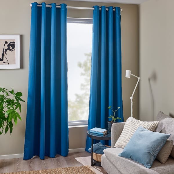 Salon avec rideaux occultants bleu foncé, œillets en acier inoxydable sur une large fenêtre, à côté d’un fauteuil gris avec oreiller bleu clair.