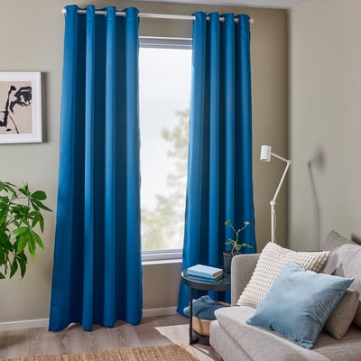 Salon avec rideaux occultants bleu foncé, œillets en acier inoxydable sur une large fenêtre, à côté d’un fauteuil gris avec oreiller bleu clair.