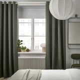 Chambre moderne avec rideaux escamotables MAJGULL vert foncé, œillets en acier inoxydable et literie blanche. Grande suspension ronde ; Vue sur la ville à l’extérieur.