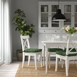 Un coin repas avec des chaises blanches et des coussins verts. Une grande plante est assise sur une chaise, mettant en valeur la taille et la couleur des coussins par rapport aux autres articles.