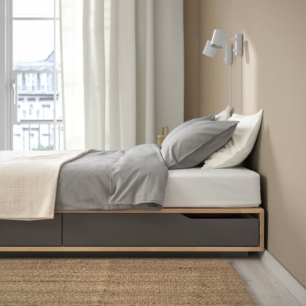 Lit avec de grands tiroirs de rangement sous matelas, doté d’un cadre en bois robuste et d’un sommier à lattes, positionné près d’une fenêtre avec des rideaux beiges.