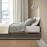 Lit avec de grands tiroirs de rangement sous matelas, doté d’un cadre en bois robuste et d’un sommier à lattes, positionné près d’une fenêtre avec des rideaux beiges.
