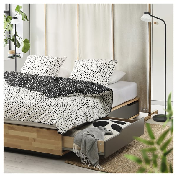 Chambre moderne avec lit en bois MANDAL, literie à pois noirs et blancs, rangement sous le lit, lampadaire et plantes.