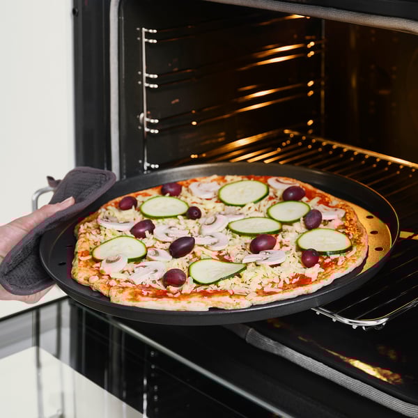 Tenant à la main un moule à pizza avec une pizza garnie de fromage, de courgettes et d’olives dans un four ouvert.