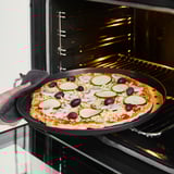 Tenant à la main un moule à pizza avec une pizza garnie de fromage, de courgettes et d’olives dans un four ouvert.