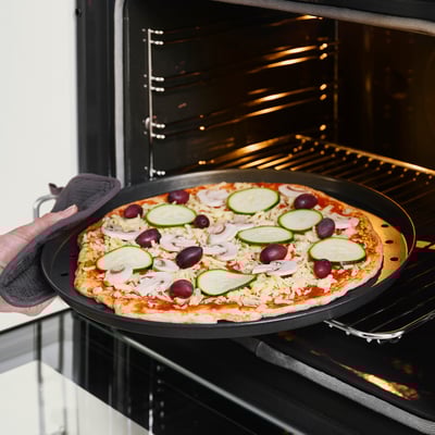 Tenant à la main un moule à pizza avec une pizza garnie de fromage, de courgettes et d’olives dans un four ouvert.