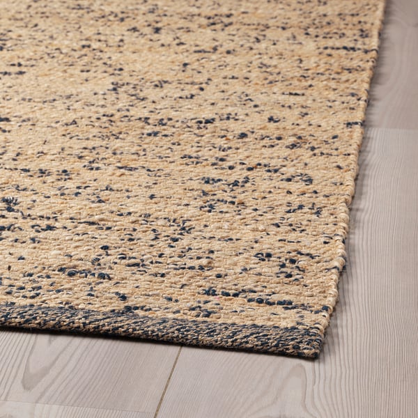 Tapis en jute naturel tissé à la main avec des taches de laine foncée aléatoires, exposé sur un parquet en bois clair.