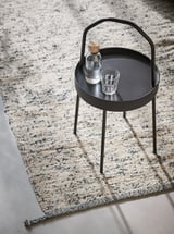 Petite table plateau ronde noire sur un tapis tressé multicolore. Il contient un verre et une bouteille.