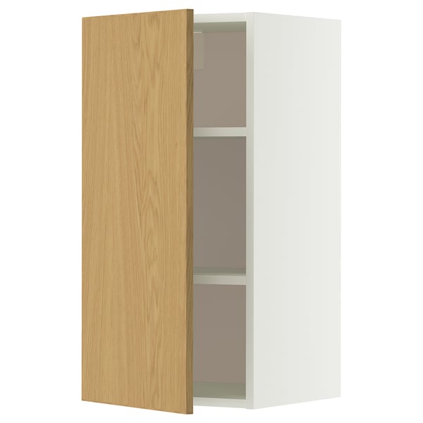 Une armoire murale METOD blanche avec une porte en chêne clair s’ouvre, révélant deux étagères en verre à l’intérieur.