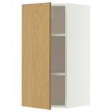 Une armoire murale METOD blanche avec une porte en chêne clair s’ouvre, révélant deux étagères en verre à l’intérieur.