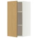 Une armoire murale METOD blanche avec une porte en chêne clair s’ouvre, révélant deux étagères en verre à l’intérieur.
