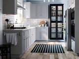 Cuisine moderne avec armoires grises, appareils électroménagers en acier inoxydable, tapis à rayures noires et blanches. L’éclairage LED sous les armoires met en valeur les plans de travail.
