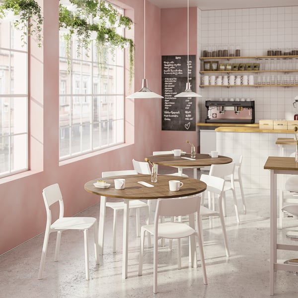 Café moderne avec des tables rondes en bois et des chaises blanches, des murs roses, de grandes fenêtres, des plantes suspendues et un comptoir à café avec des étagères.
