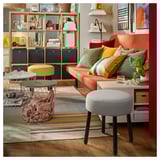 Salon coloré : pouf gris, canapé orange, tabouret jaune, tapis multicolore, étagères en bois avec décor.