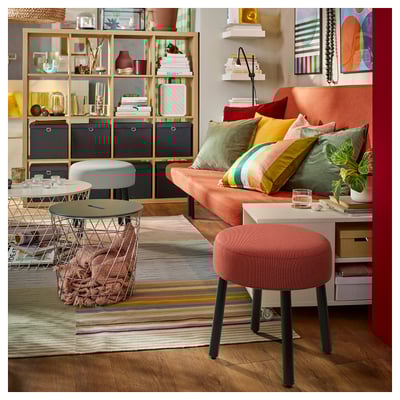 Salon lumineux avec canapé orange, oreillers colorés, tapis rayé, meubles en fil de fer et plantes. Tabouret orange centré.