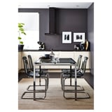 Salle à manger moderne avec table en verre, chaises transparentes, accents noirs, murs gris, tapis, plantes, art et appareils de cuisine.