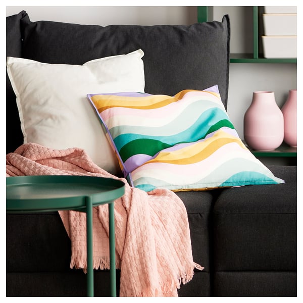 Coussin aux couleurs vives avec des rayures ondulées sur un canapé noir. Accompagné d’une couverture rose clair et de vases.