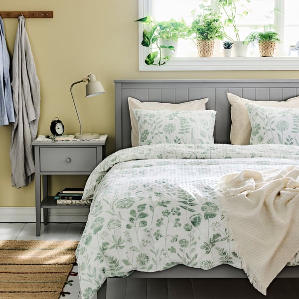 Chambre douillette avec une couette fleurie verte et blanche en NÅLBJÖRNBÄR , un cadre de lit en bois, une table de chevet grise, un mur jaune et des plantes.