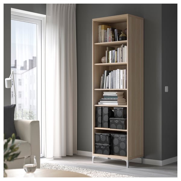 Une étagère en bois clair dans une pièce. Il contient divers livres et bacs de rangement. Un rideau blanc et une chaise se trouvent à proximité.