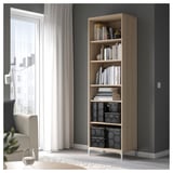 Une étagère en bois clair dans une pièce. Il contient divers livres et bacs de rangement. Un rideau blanc et une chaise se trouvent à proximité.