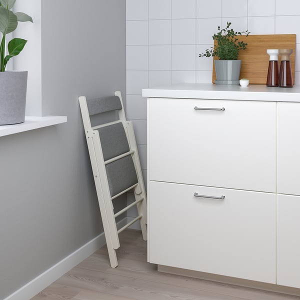 Petite table de cuisine blanche avec quatre tiroirs, chaises pliantes rembourrées grises, comptoir en bois avec poignée, mur carrelé gris, plancher en bois.