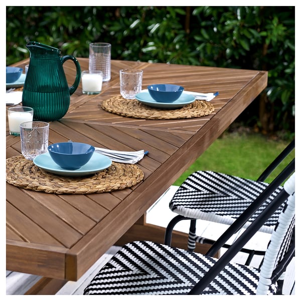 Salle à manger en plein air avec table en bois norrmanso, chaises en osier, bols bleus, verres et pichet vert.
