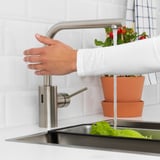 Personne se lave les mains sous un robinet en acier inoxydable avec de l’eau qui coule dans l’évier de la cuisine ; plantes en pot à proximité.