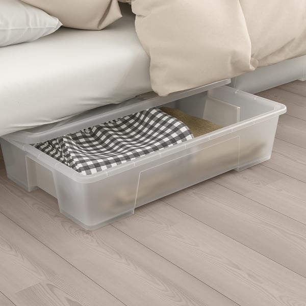 Une boîte de rangement en plastique transparent sous un lit, remplie de couvertures pliées, démontrant une utilisation efficace de l’espace de stockage.