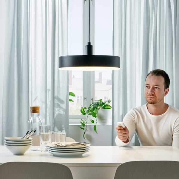 Homme avec lampe NYMÅNE, télécommande, table dressée, plante par fenêtre.
