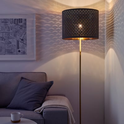 Un salon moderne avec un lampadaire NYMÖ projetant une lumière à motifs sur le mur. La lampe a une base de couleur laiton et un abat-jour perforé noir.