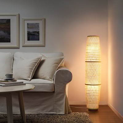 Lampadaire ÖGONGULL avec abat-jour en bambou à côté du canapé beige, émettant une lueur chaude.