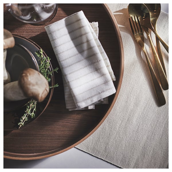 Un élégant plateau de service OMBONAD en bois sombre avec des couverts dorés, des serviettes à rayures blanches et un petit plat avec des herbes.