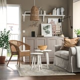 Salon moderne avec tapis bleu clair ONSEVIG, chaises en osier, canapé gris, tables d’appoint blanches et armoire en bois avec décorations.