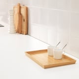 Plateau en bois avec deux tasses et cuillères blanches, posé sur un comptoir blanc à côté de planches à découper en bois et d’une pile d’assiettes blanches.