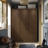 Armoire PAX moderne en bois brun foncé, haute et large, avec des poignées simples, entre les fenêtres et les crochets muraux, sur tapis gris.