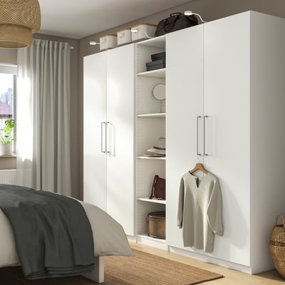 Une armoire PAX blanche dans une chambre. Il est plus grand que le lit et a des étagères ouvertes avec des vêtements et des accessoires. Un pull gris est suspendu sur le devant.