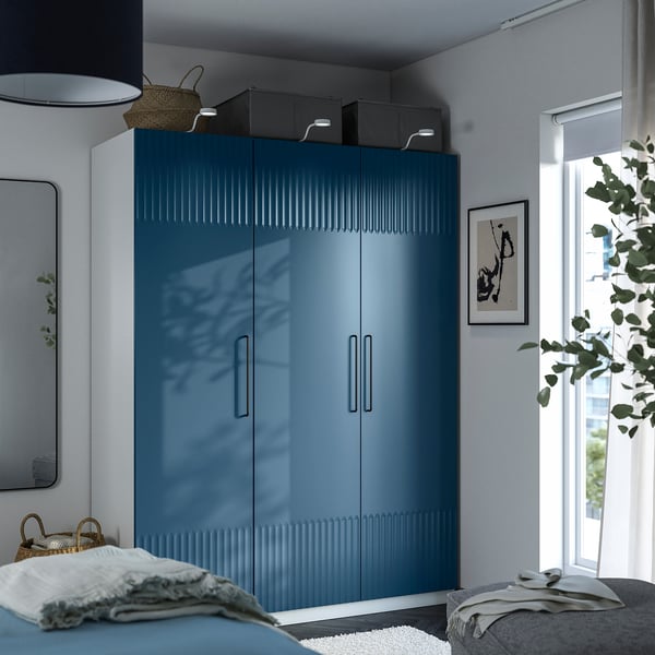 Chambre moderne avec grande armoire PAX bleue, poignées argentées, lumière naturelle, plante, linge de lit clair