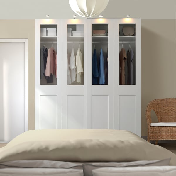 Armoire PAX blanche avec des vêtements colorés, grand lit et chaise en osier.
