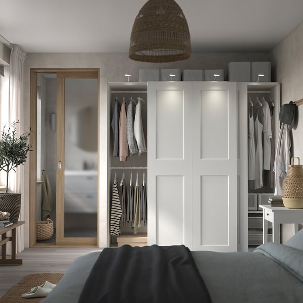 Chambre moderne avec armoire PAX blanche, vêtements ouverts, miroir, lampe, plante.