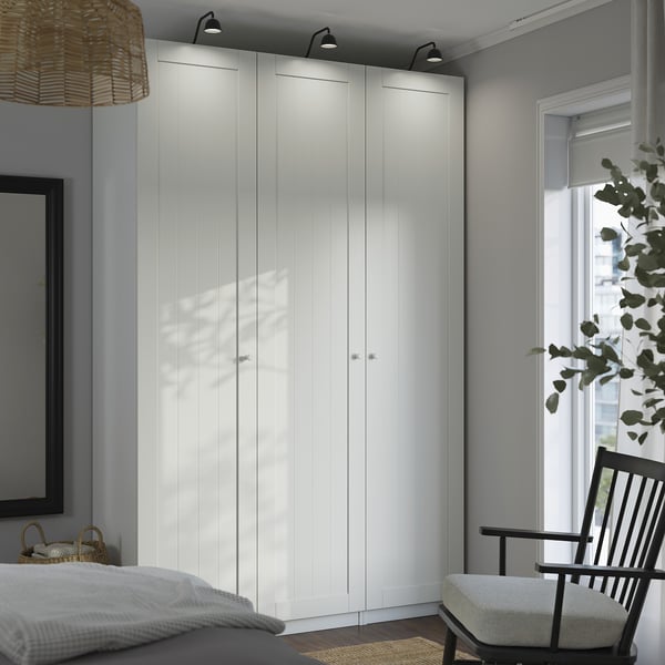 Chambre minimaliste moderne avec grande armoire PAX blanche, mur haut et étendu, à côté d’une fenêtre à cadre noir et d’une chaise en bois.
